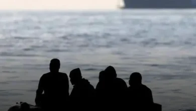 Four people silhouetted on a small boat at sea with a large ship in the background
